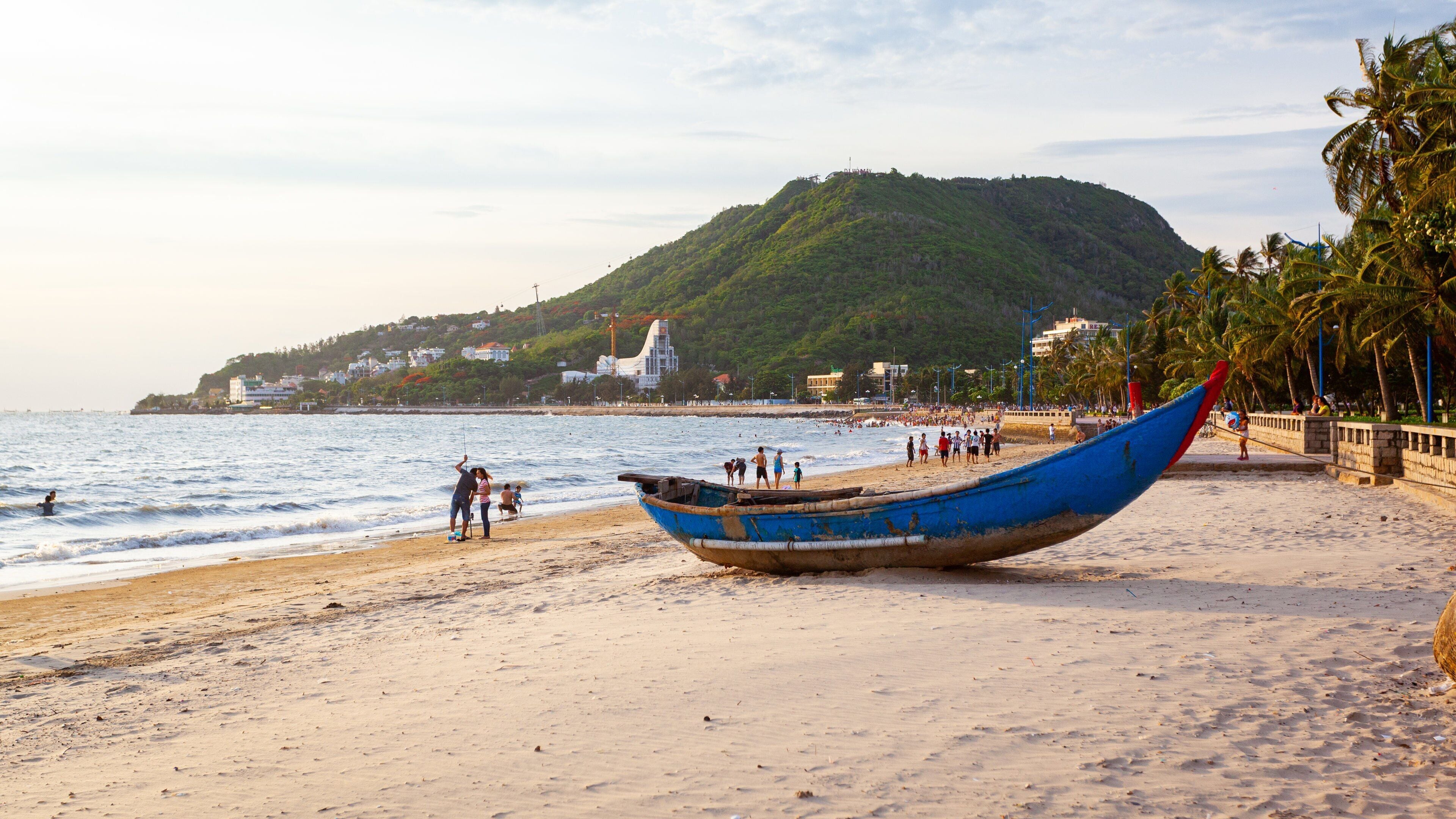 A beach with a large blue boat, a sandy shore, and a hill with buildings in the background.