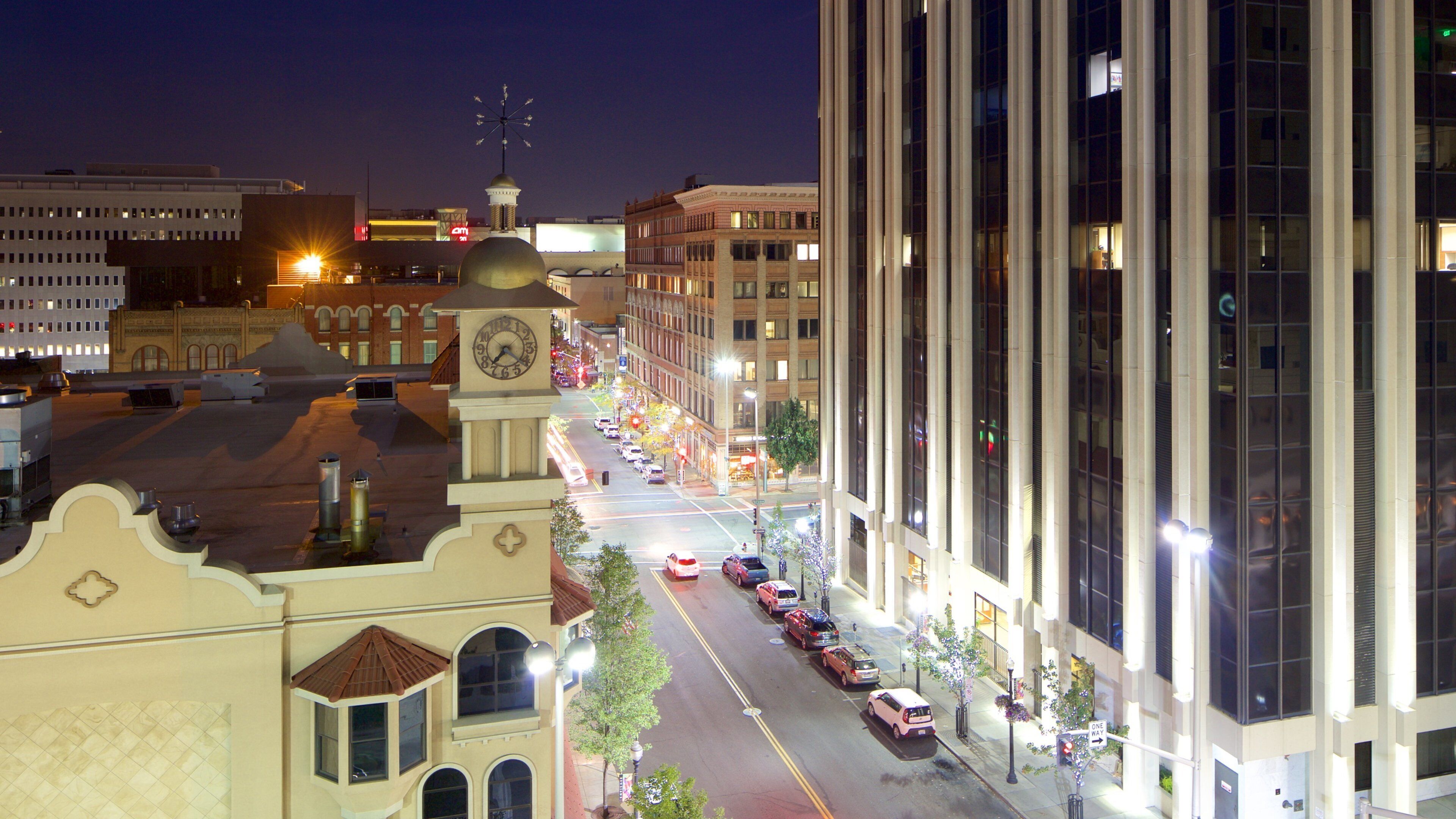 A nighttime cityscape with a prominent clock tower, a well-lit street lined with parked cars, and modern buildings with reflective windows.