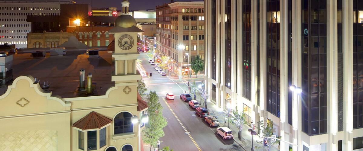 A nighttime cityscape with a prominent clock tower, a well-lit street lined with parked cars, and modern buildings with reflective windows.
