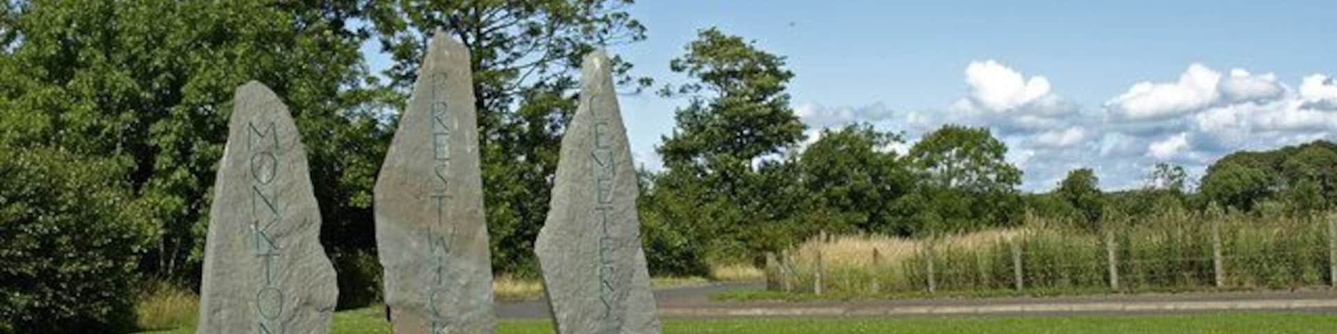 Tres piedras verticales en un campo cubierto de hierba, con árboles al fondo.