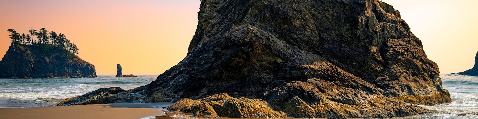 Formação rochosa com árvores no topo, uma praia de areia e o oceano ao fundo.