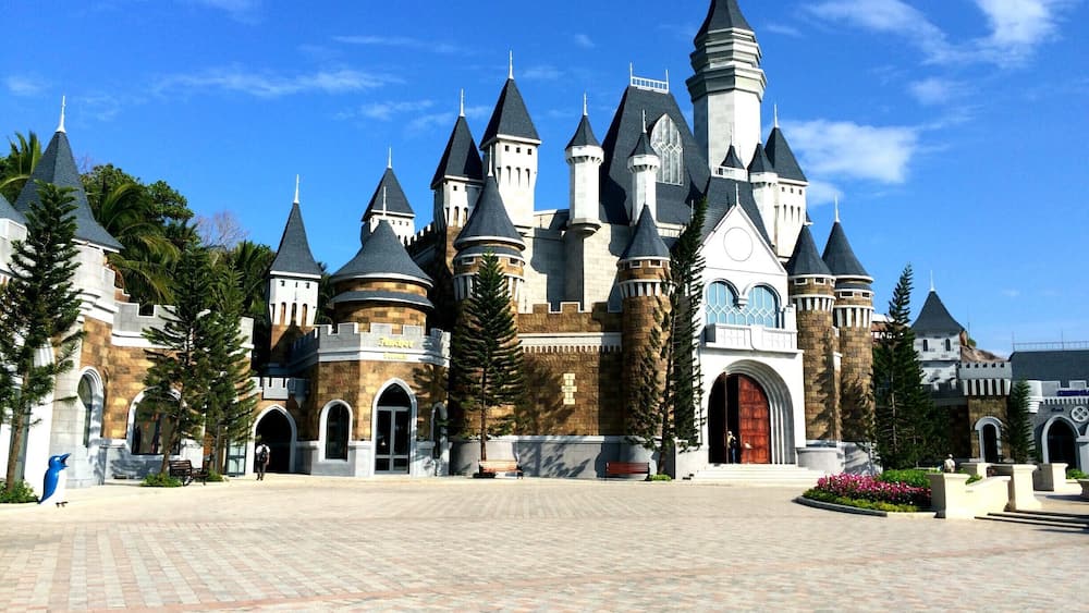 A large castle-like building with multiple towers and turrets, surrounded by a paved courtyard and palm trees.