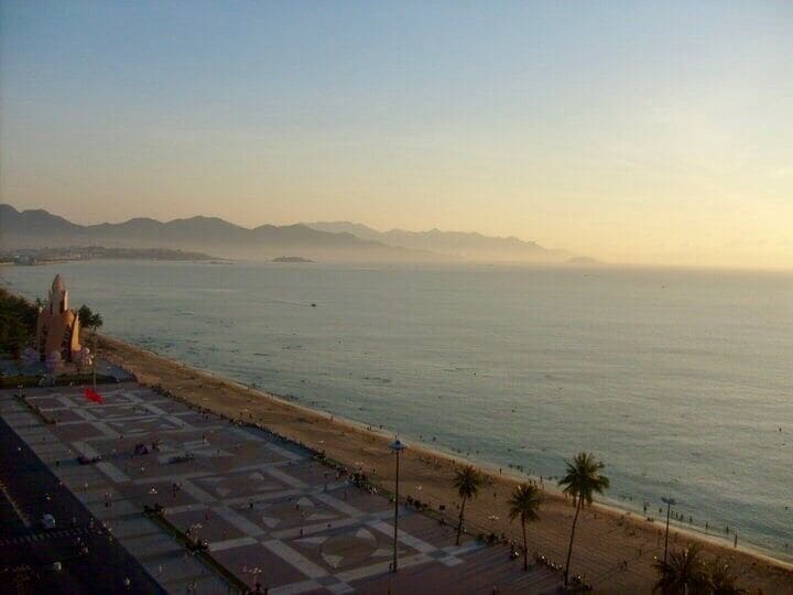 Vista costera con playa, palmeras y montañas a lo lejos.