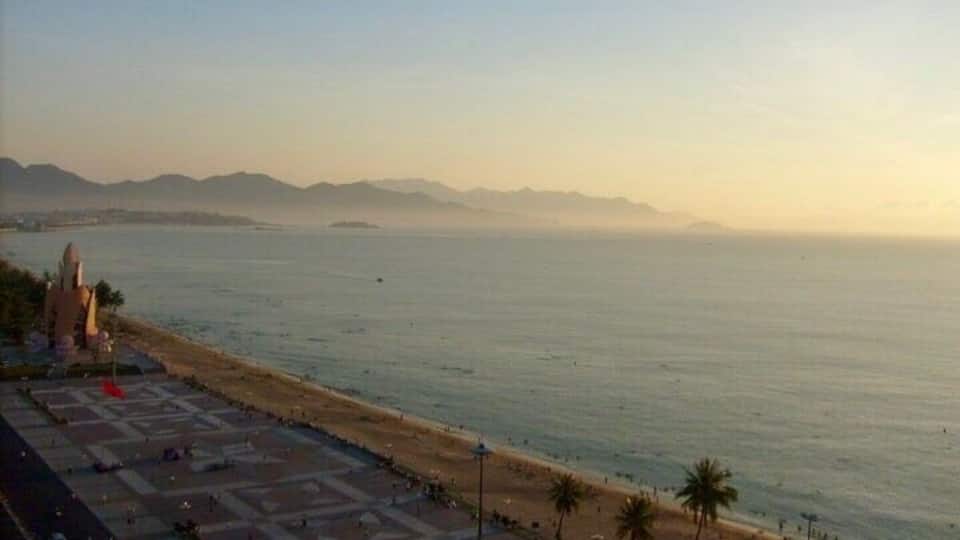Vista costera con playa, palmeras y montañas a lo lejos.