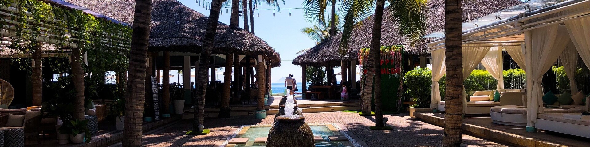 Un resort tropical con una fuente central, palmeras y construcciones de techo de paja.