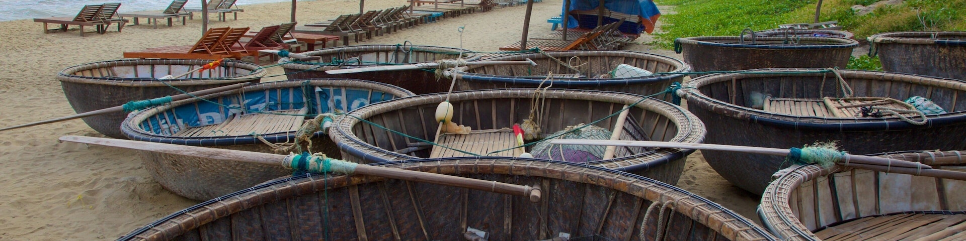 Guarda-sóis de palha, espreguiçadeiras de madeira e barcos tradicionais numa praia.