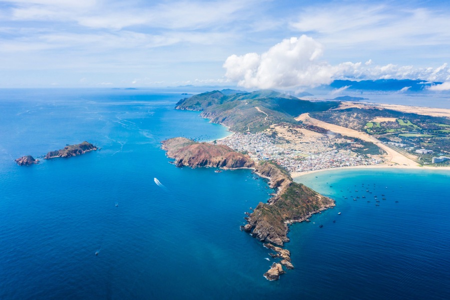 Vista aérea de un pueblo costero con playa, afloramientos rocosos y montañas al fondo.