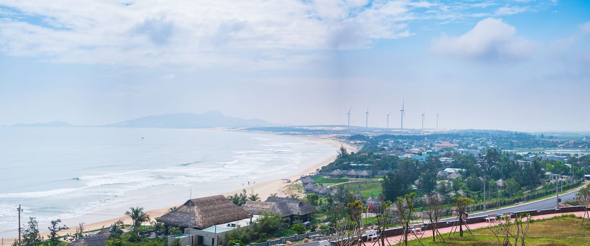 A coastal town with a beach, wind turbines, and a mountain in the distance.