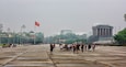 A large open plaza with a crowd of people, a prominent flag, and several buildings in the background.