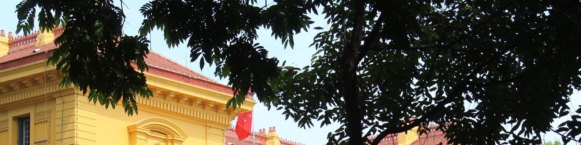 A yellow building with ornate architecture, a red flag, and a well-maintained garden.