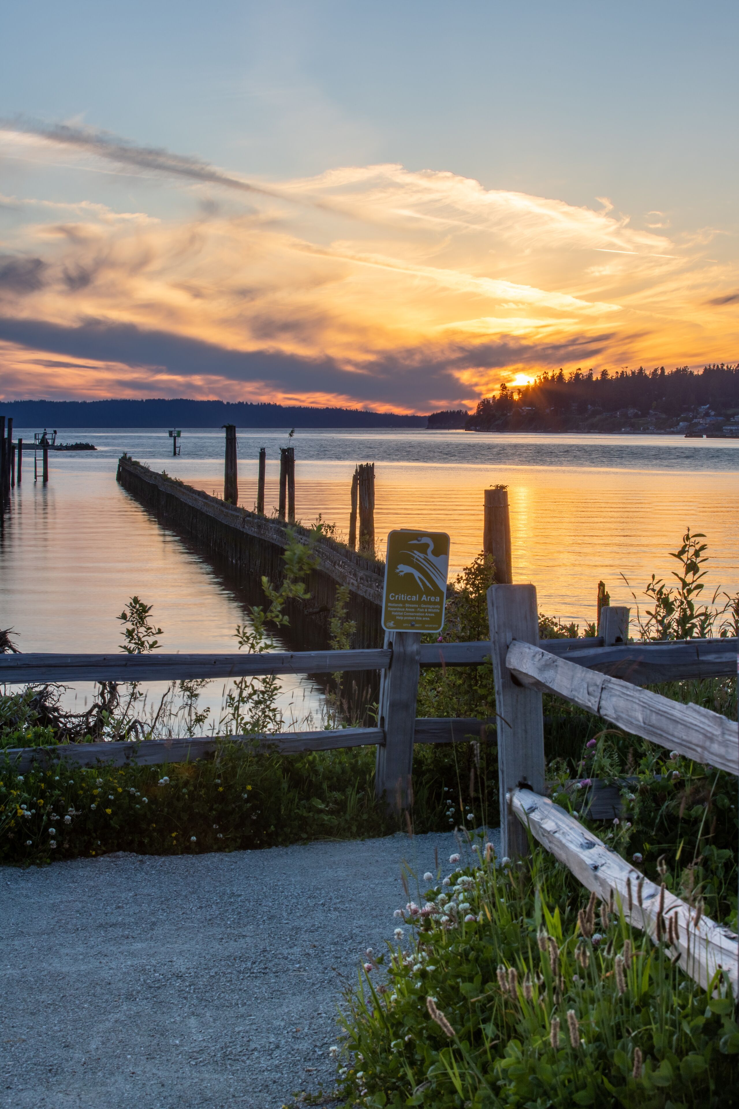 A sunset over a calm lake with a wooden pier and a sign indicating a critical area.