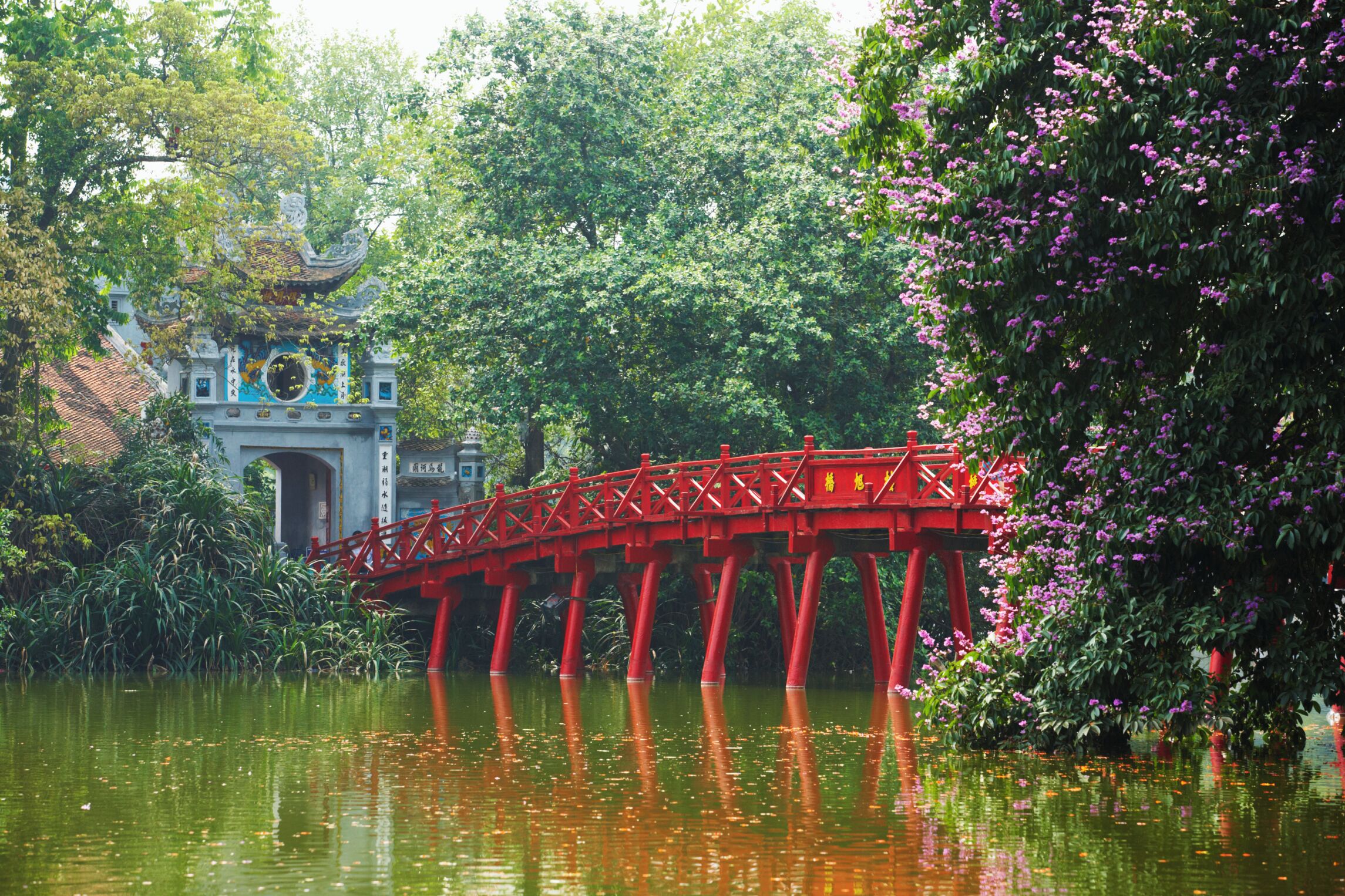 A red bridge over a calm river, surrounded by lush greenery and a traditional building with a tiled roof.