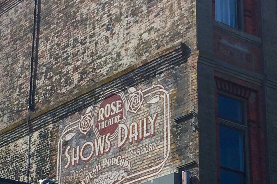 Un vieux bâtiment orné d’une enseigne rétro indiquant « Rose Theatre Shows Daily », une boutique de pop-corn et un panneau de signalisation.