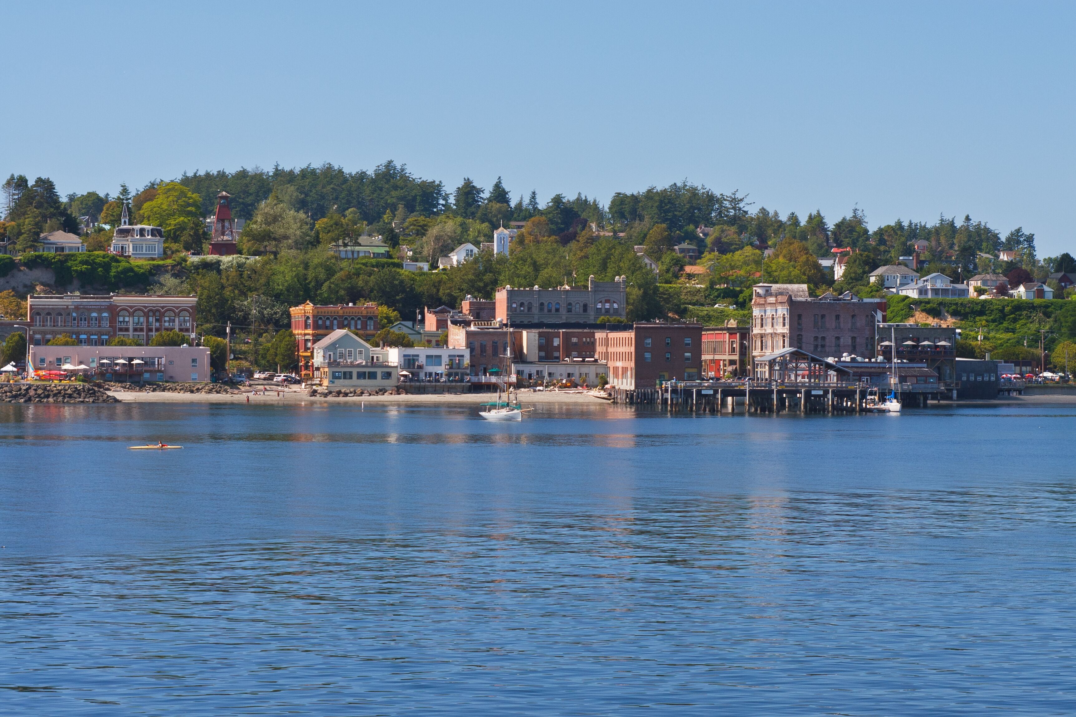 A waterfront town with historic buildings, a pier, and a hilltop with trees.