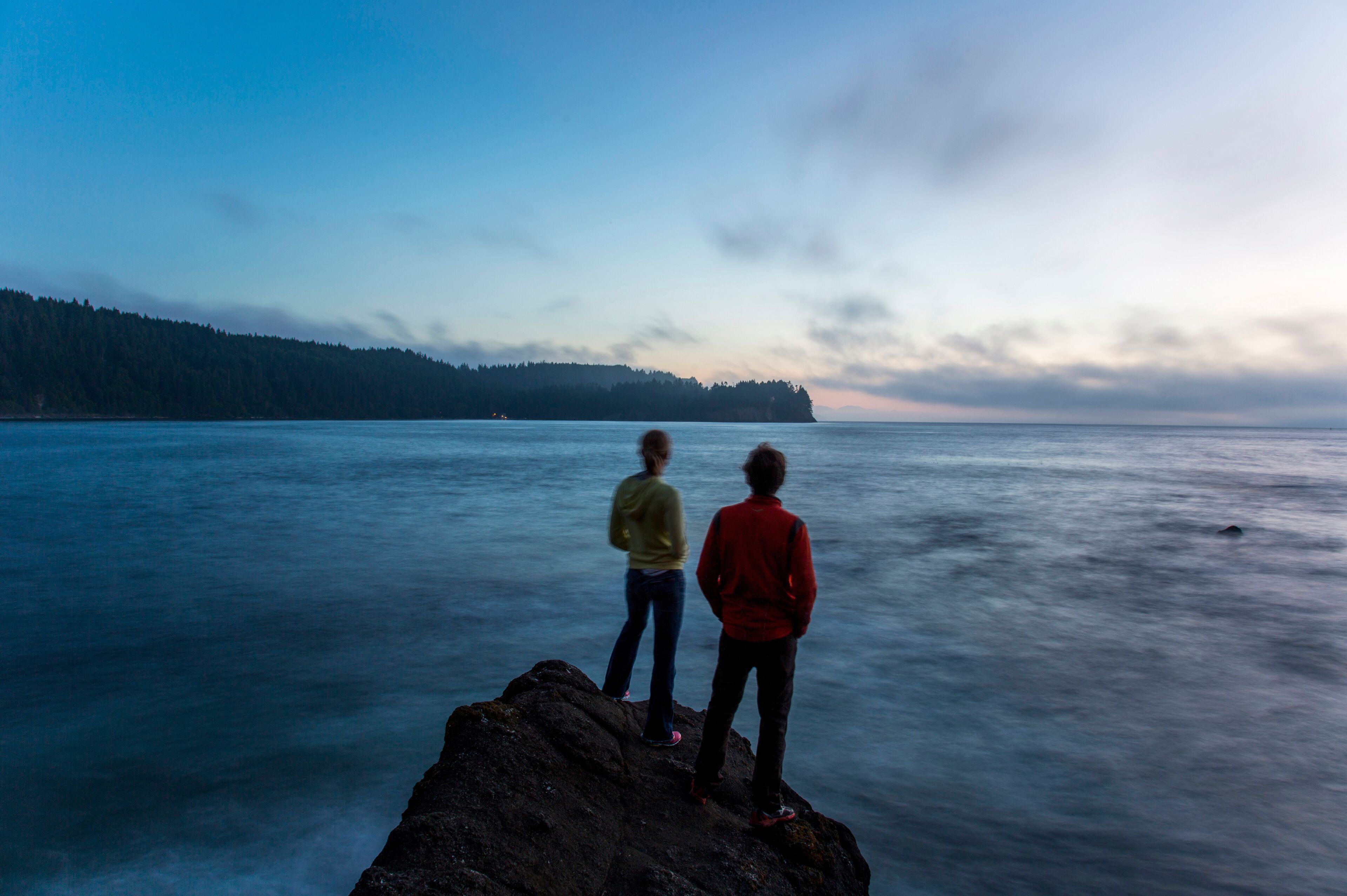 Two young people stand on the edge of a rock formation at dusk near the Strait of Juan de Fuca in Washington.