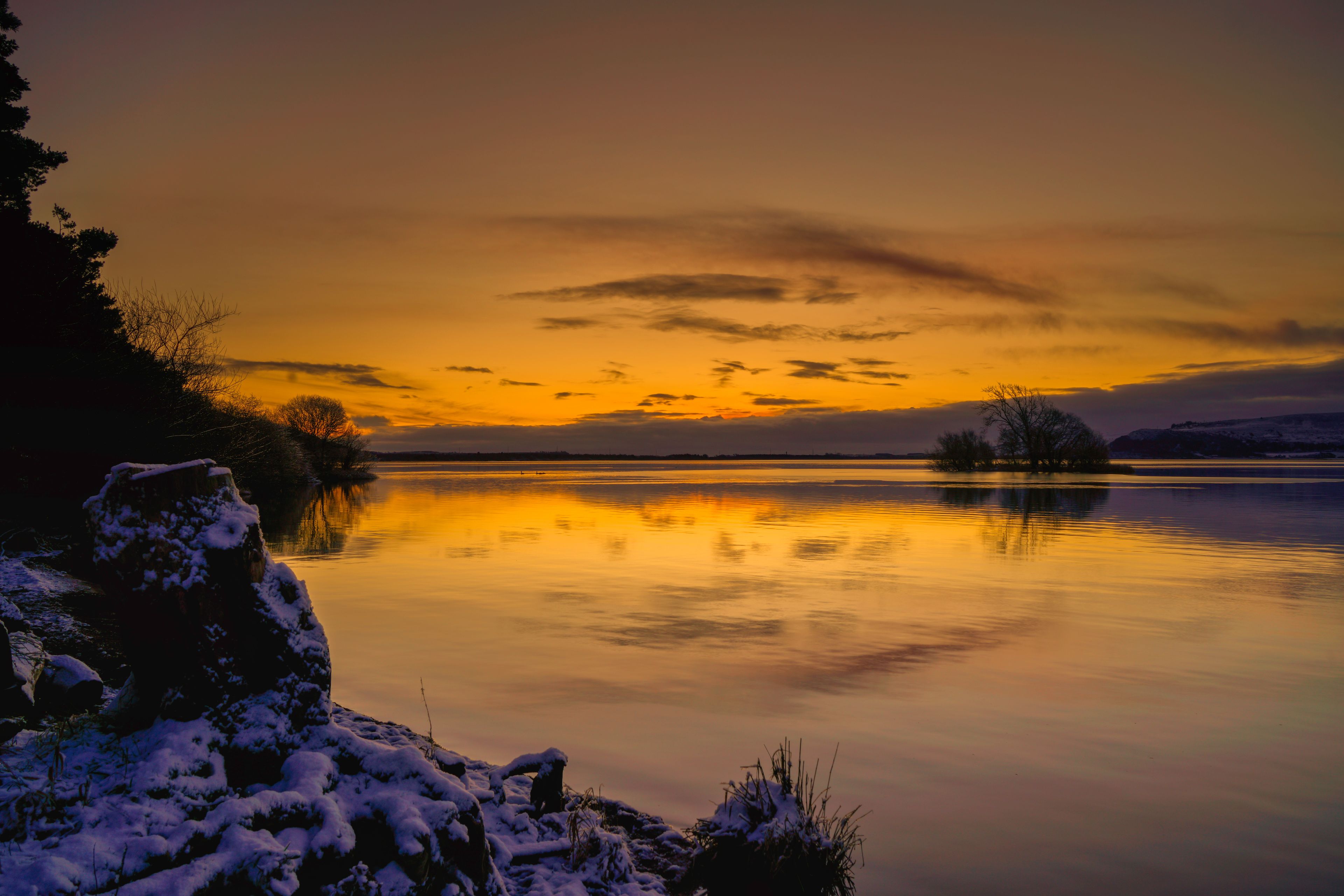 A calm lake at sunset with snow-covered rocks and trees reflecting the sky.