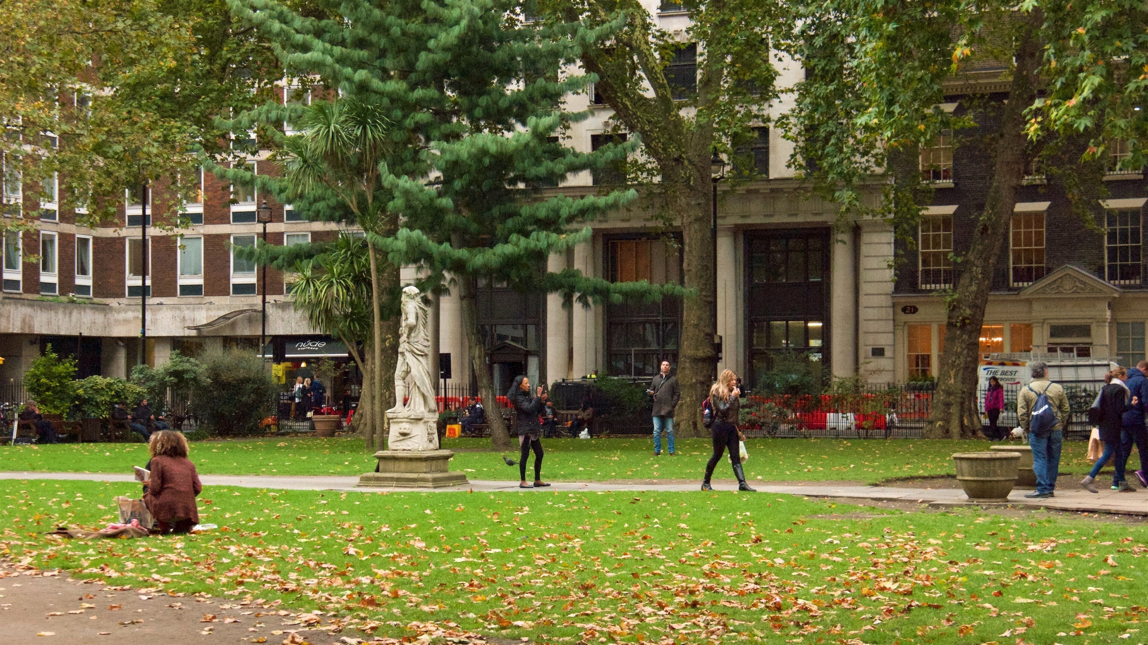 A park with a statue, people walking, and buildings in the background.