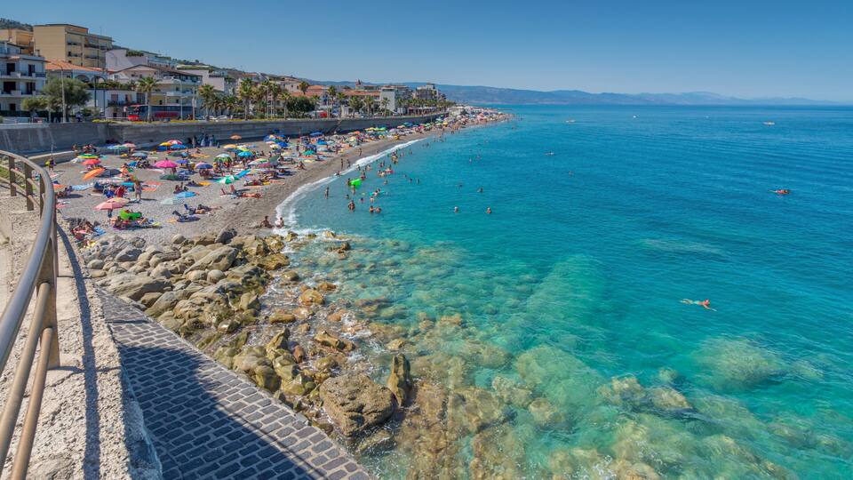A coastal beach with clear turquoise water, rocky shoreline, and buildings along the promenade.