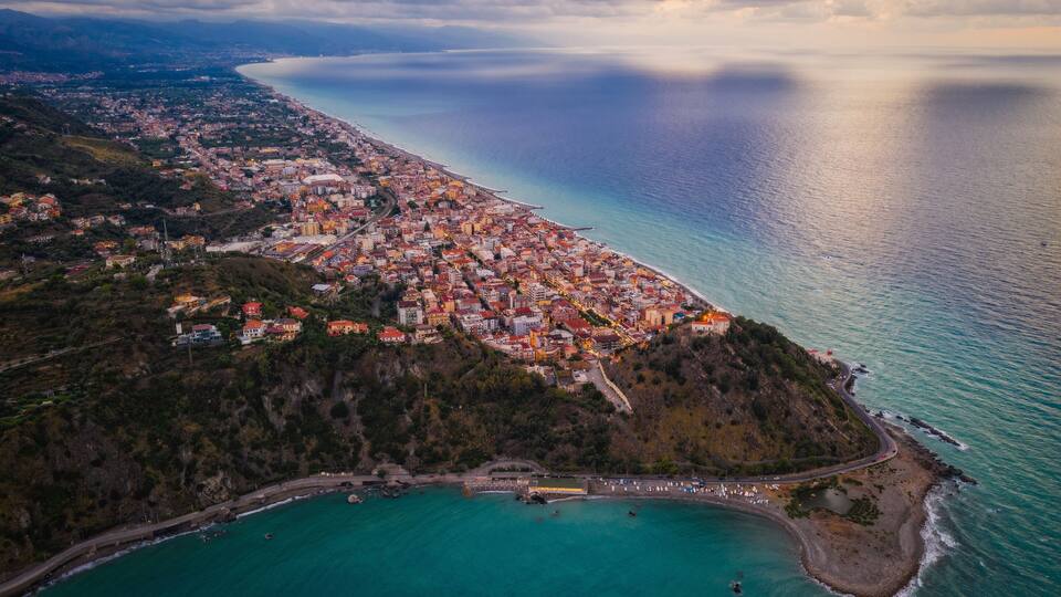 An aerial view of a coastal town with a dense cluster of buildings, a prominent hill, and a clear view of the coastline.