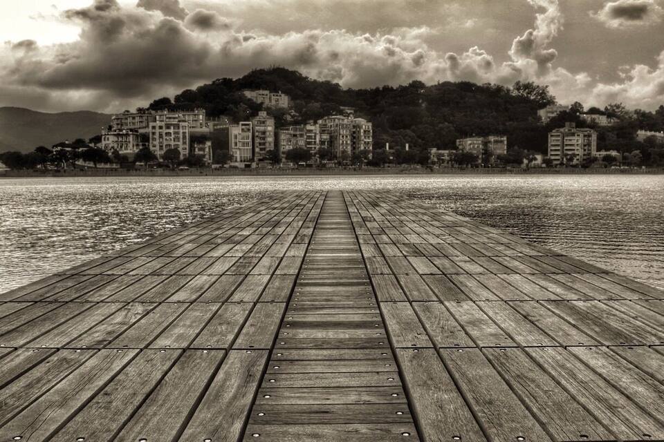 Un muelle de madera que se extiende sobre aguas tranquilas, con un paisaje urbano y colinas al fondo.