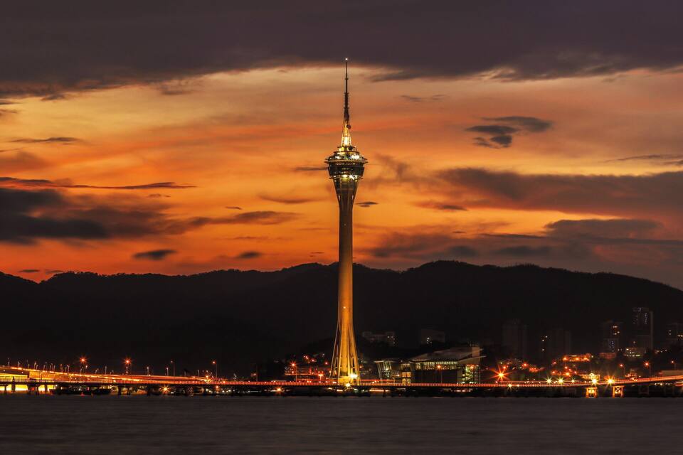 A tall tower with a spire, surrounded by a cityscape and mountains under a sunset sky.