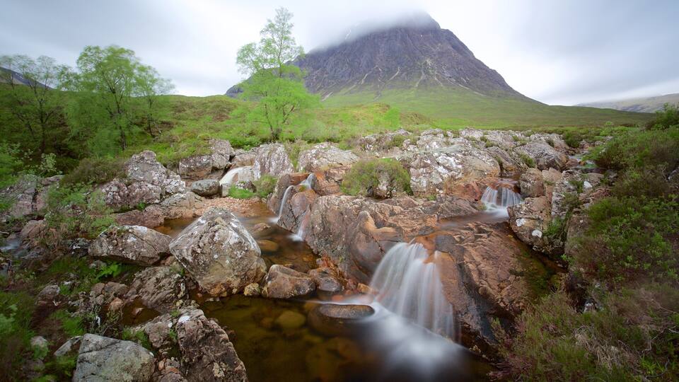 A waterfall cascading over rocks in a lush, green valley with a mountain in the background.