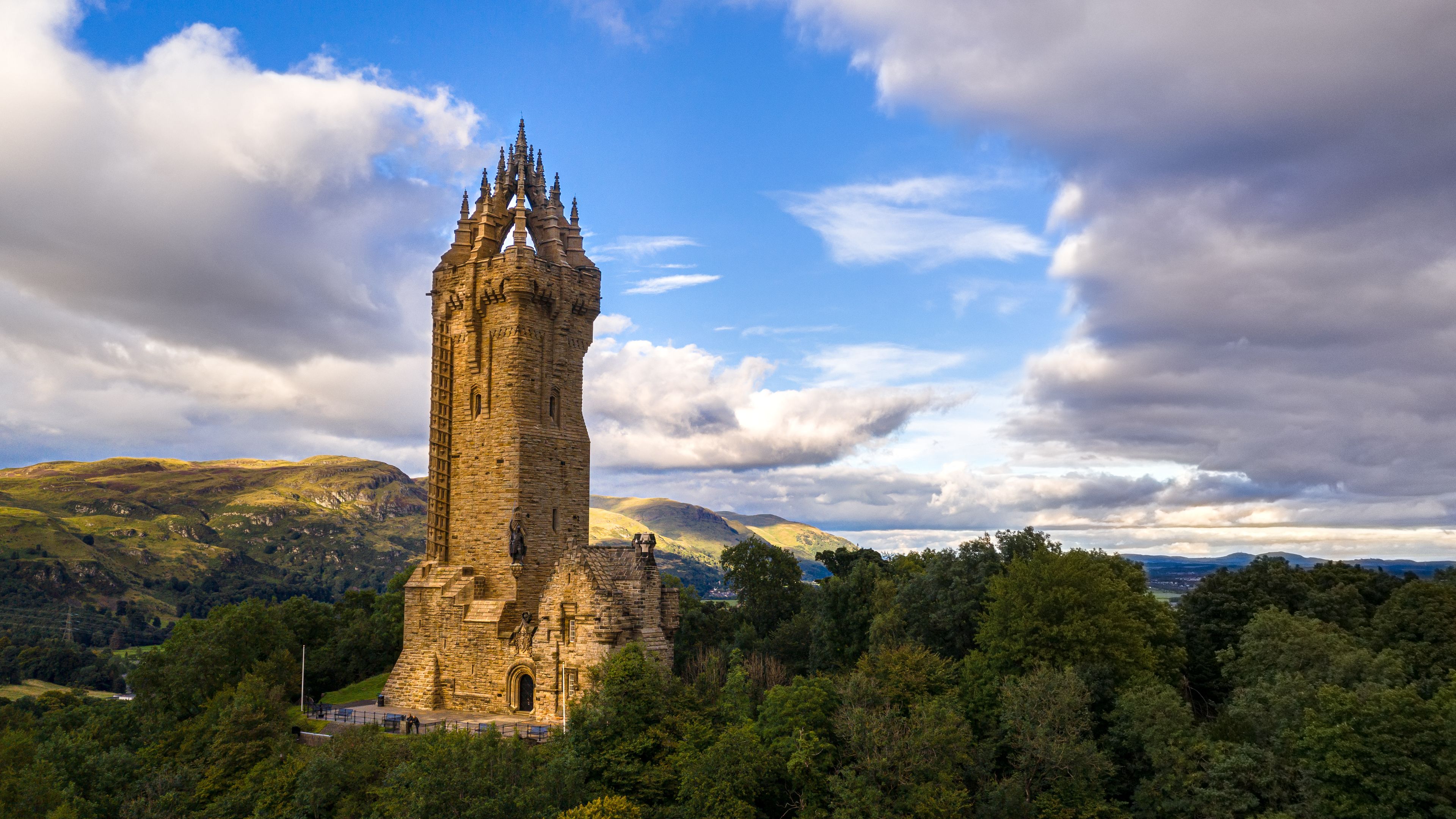 A historic stone tower with a pointed roof, surrounded by lush green hills and a partly cloudy sky.