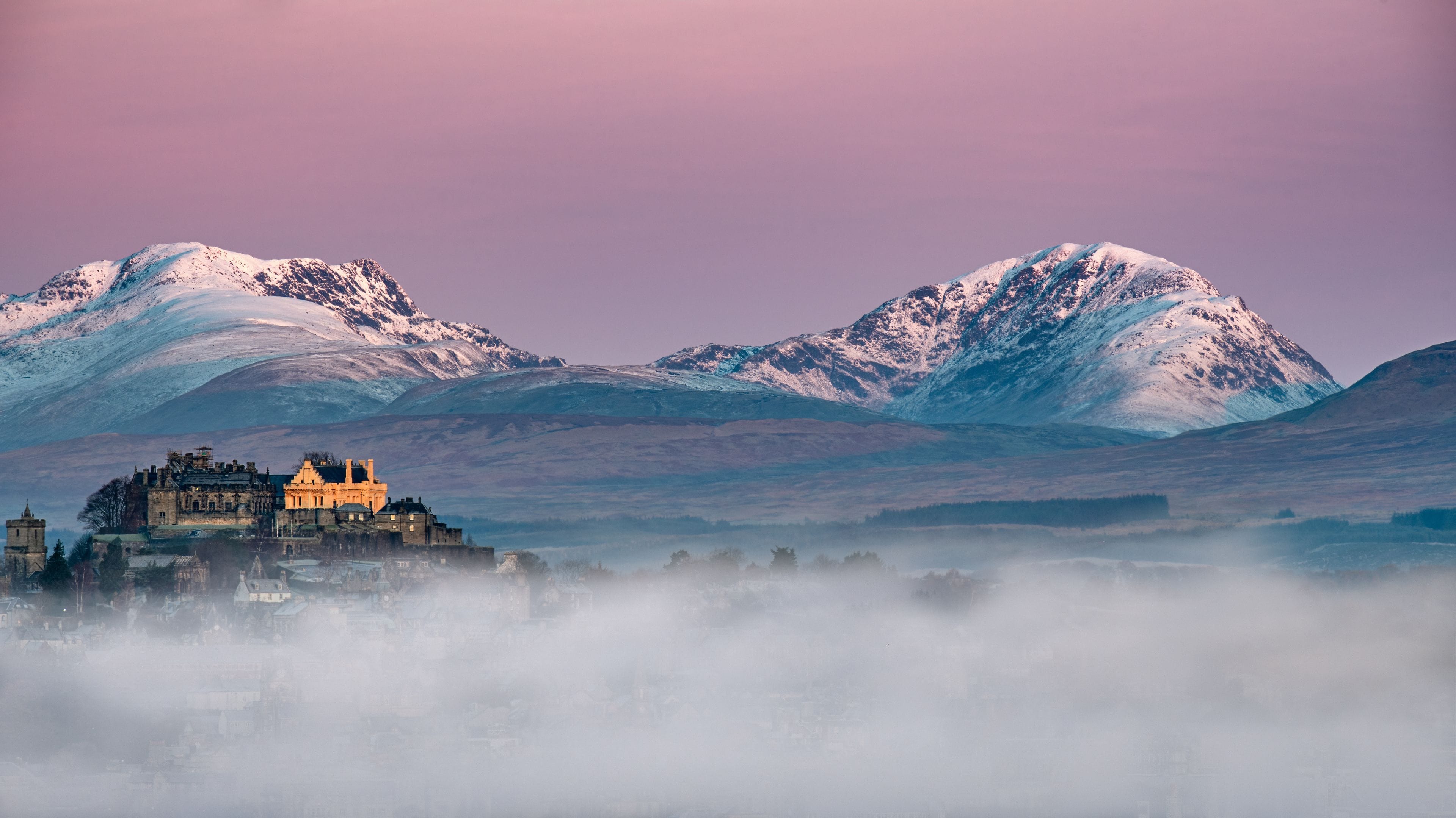A castle surrounded by snow-covered mountains and a misty foreground.