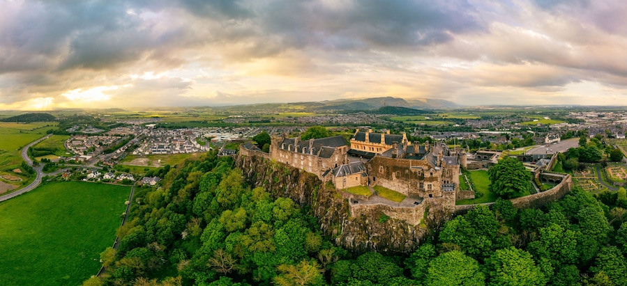 A historic castle surrounded by lush greenery, overlooking a town with a river and distant hills.