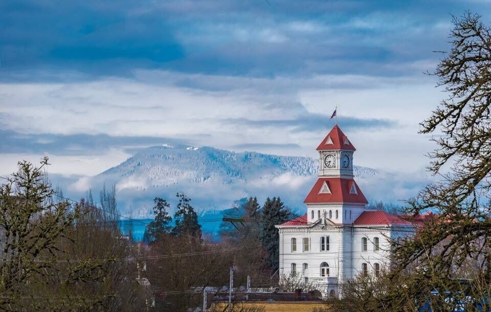 A white building with a red roof and a clock tower, surrounded by trees and mountains in the background.
