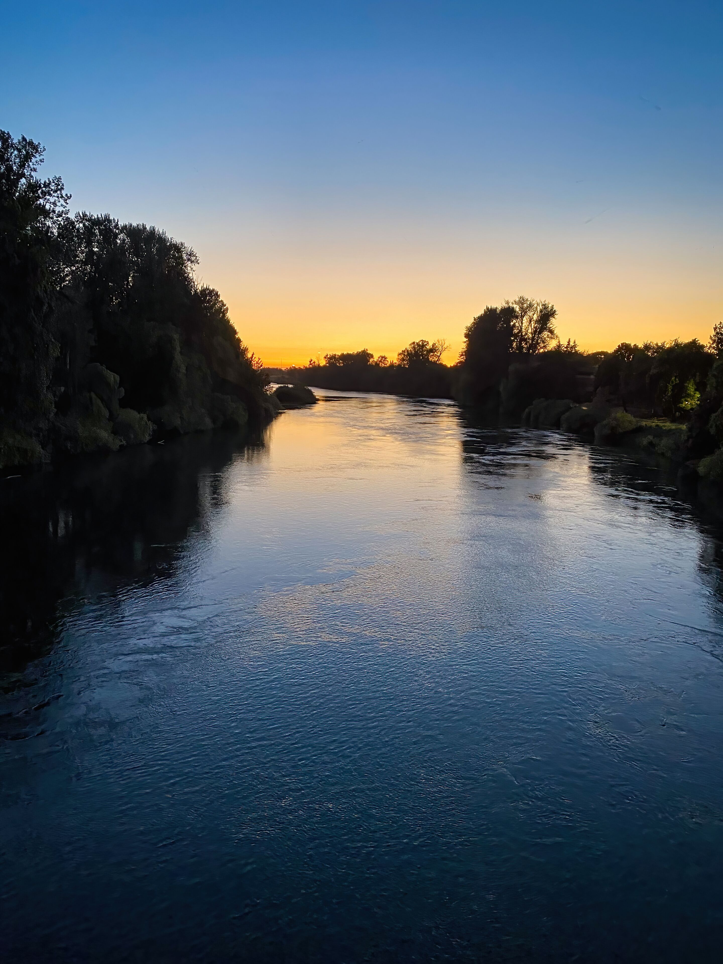 A calm river at sunset with surrounding trees and a clear sky.