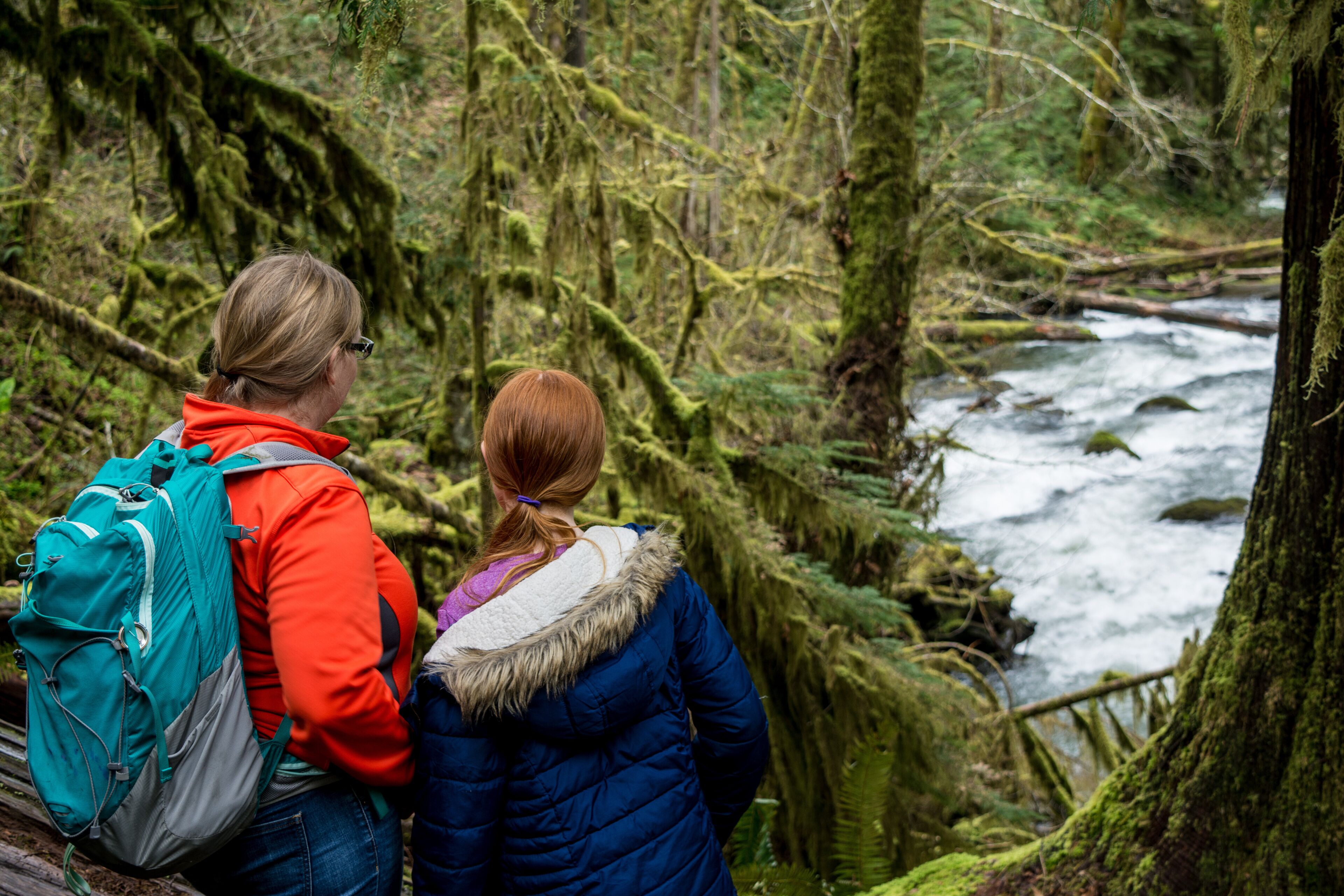 Two people standing by a forested river.