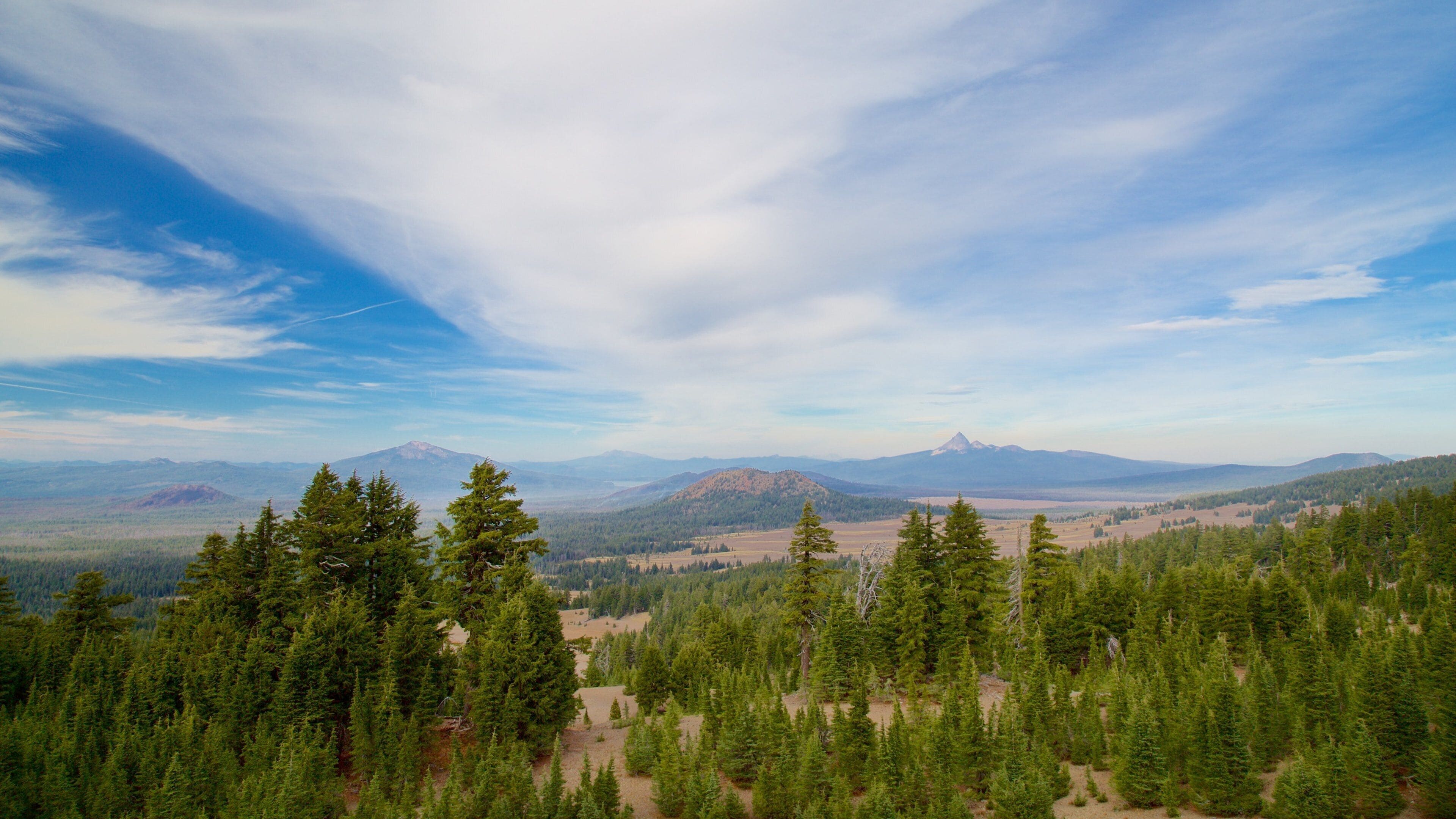 A mountainous landscape with dense forests and distant peaks under a partly cloudy sky.