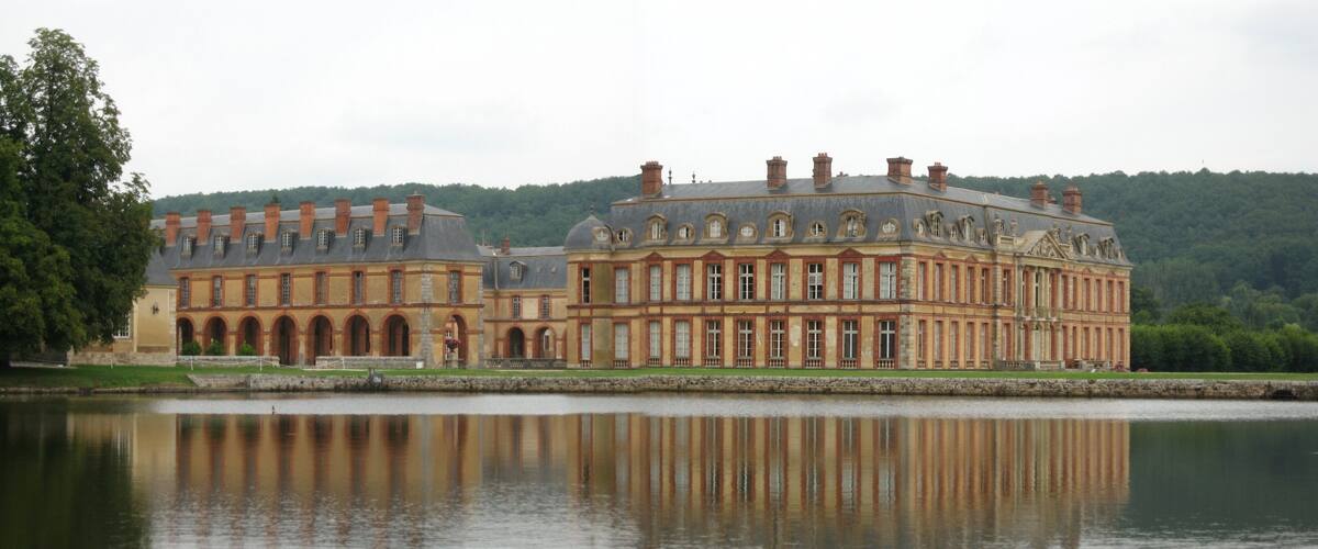 A large, historic building with a reflection in a calm body of water, surrounded by greenery and hills.