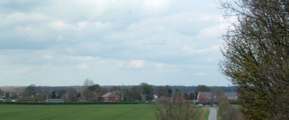 A rural road flanked by fields and trees, leading towards a distant village.