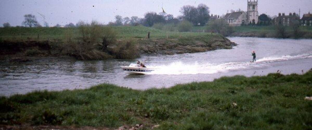 A river with a boat and a person wakeboarding. There is a building in the background.
