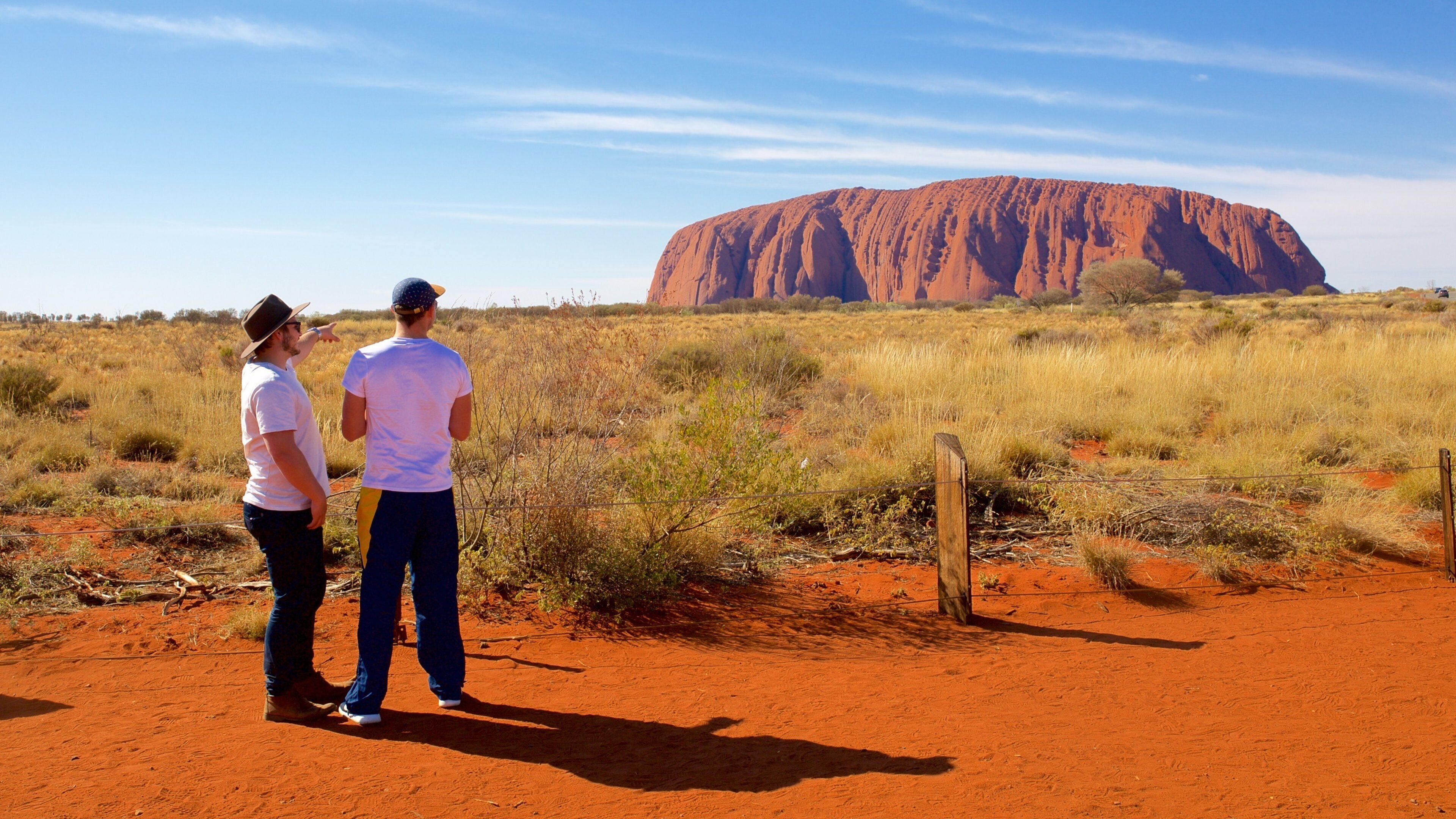 Two people standing on red soil, looking at Uluru.