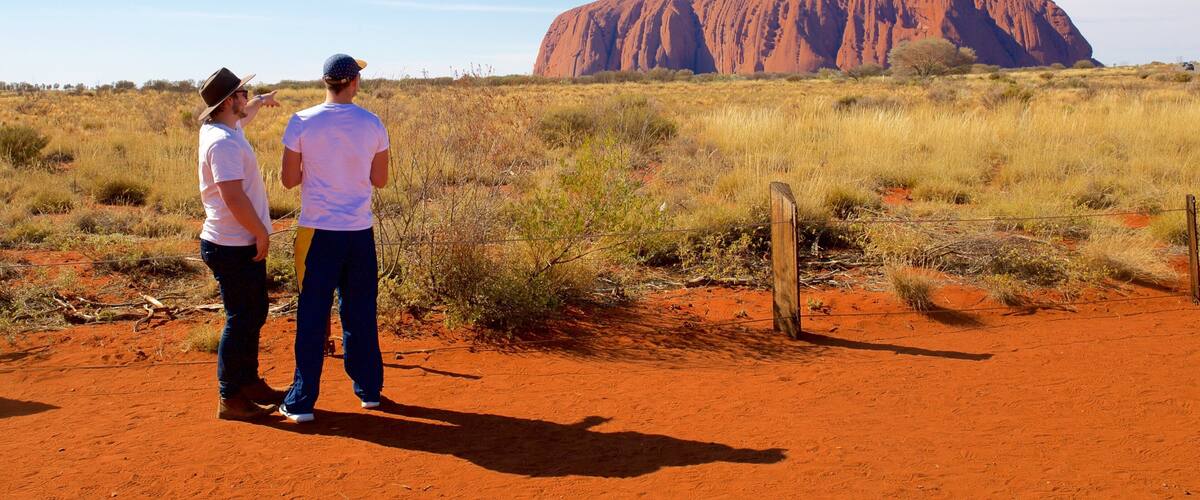 Two people standing on red soil, looking at Uluru.