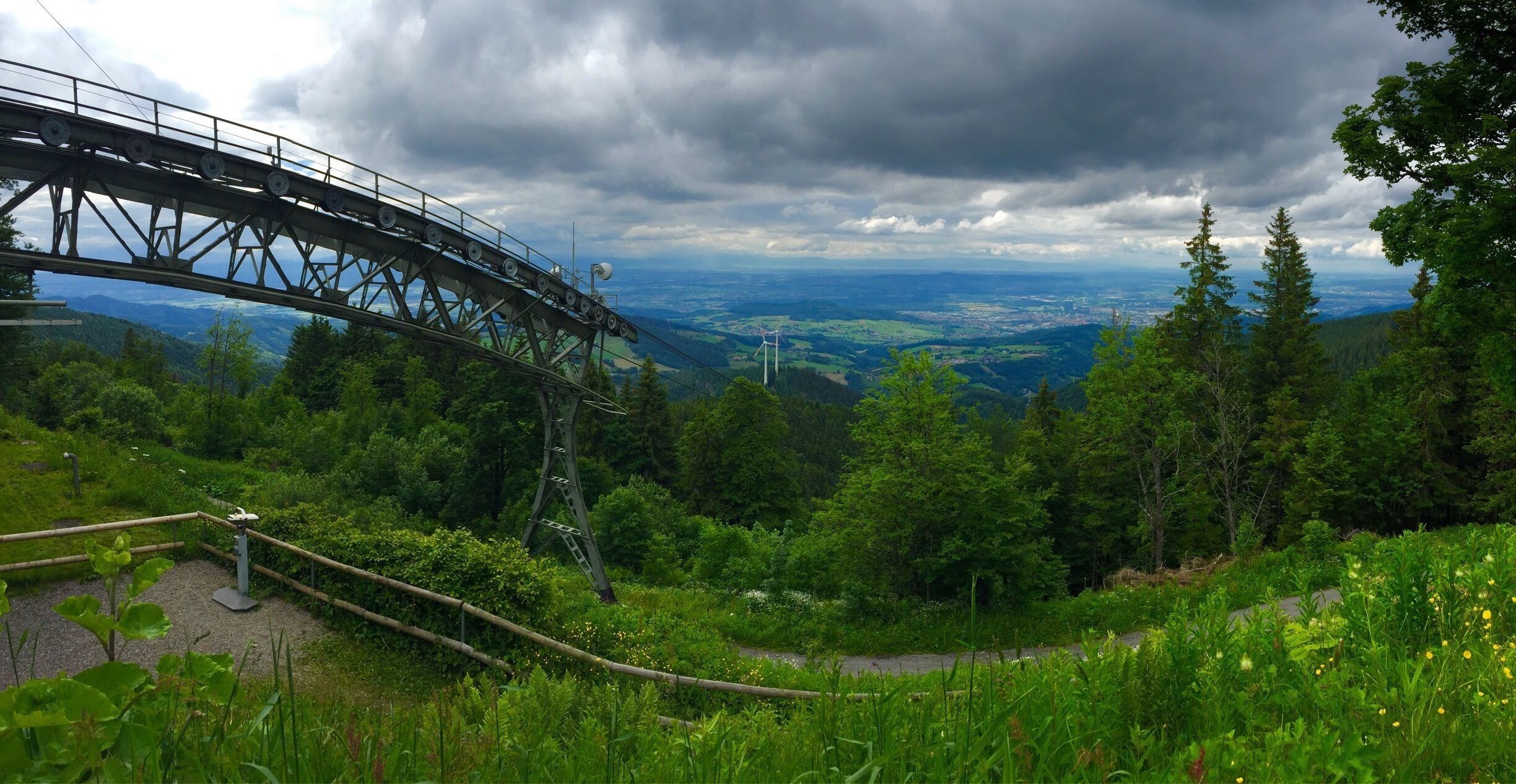 A metal bridge with a scenic view of a forested valley and distant wind turbines.