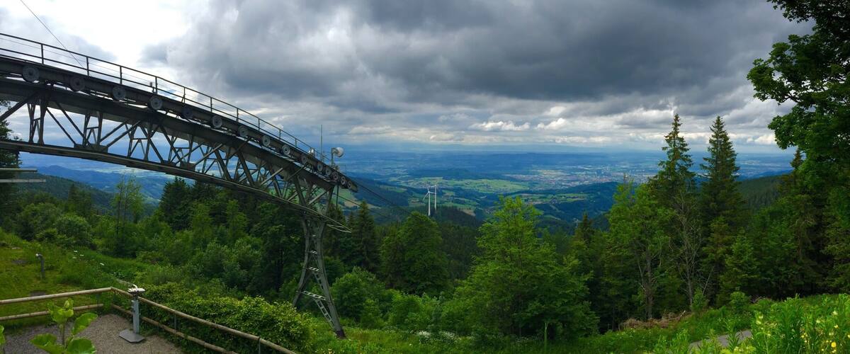 A metal bridge with a scenic view of a forested valley and distant wind turbines.