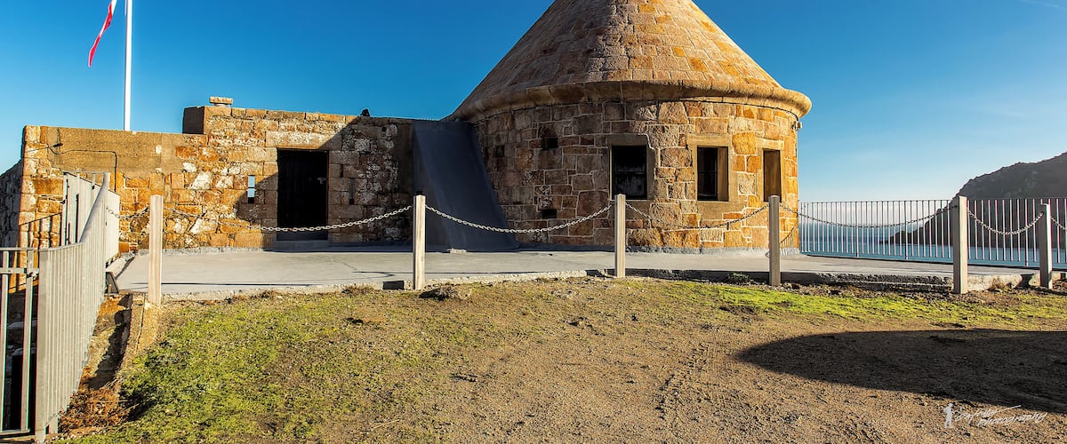 A stone building with a conical roof, a flagpole, and a chain-link fence.