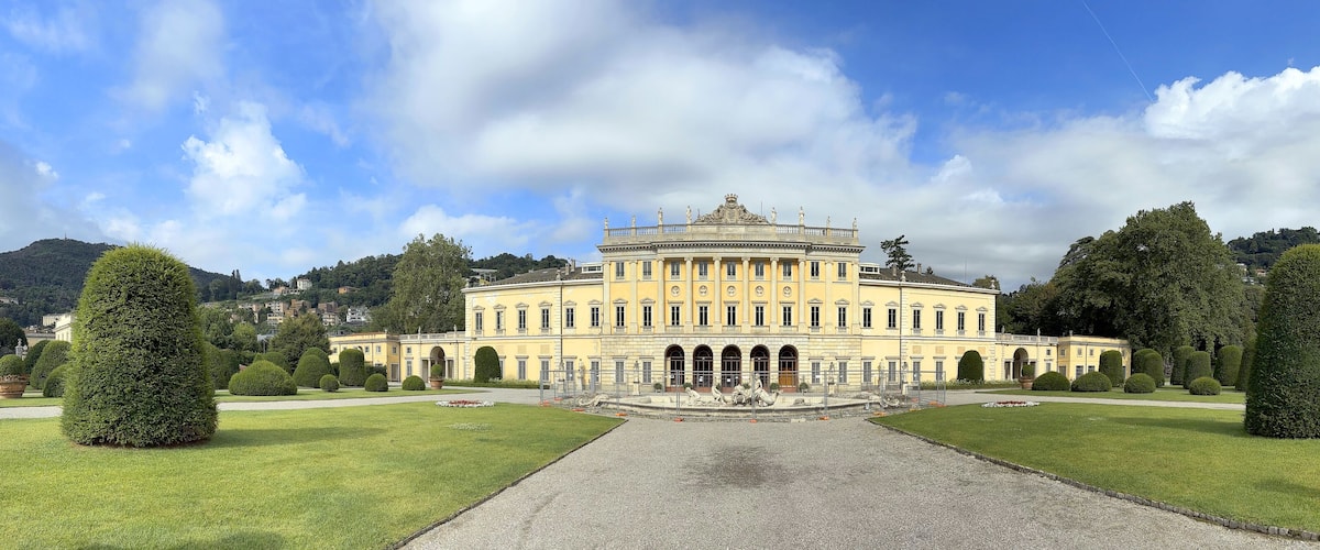 A grand yellow building with a central courtyard, surrounded by well-manicured lawns and tall trees.