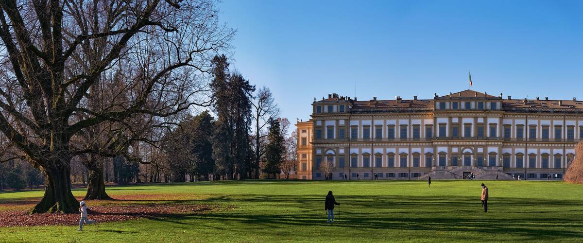 A large, classical building with a symmetrical facade and a flag on top, surrounded by a well-maintained park with grass and trees.