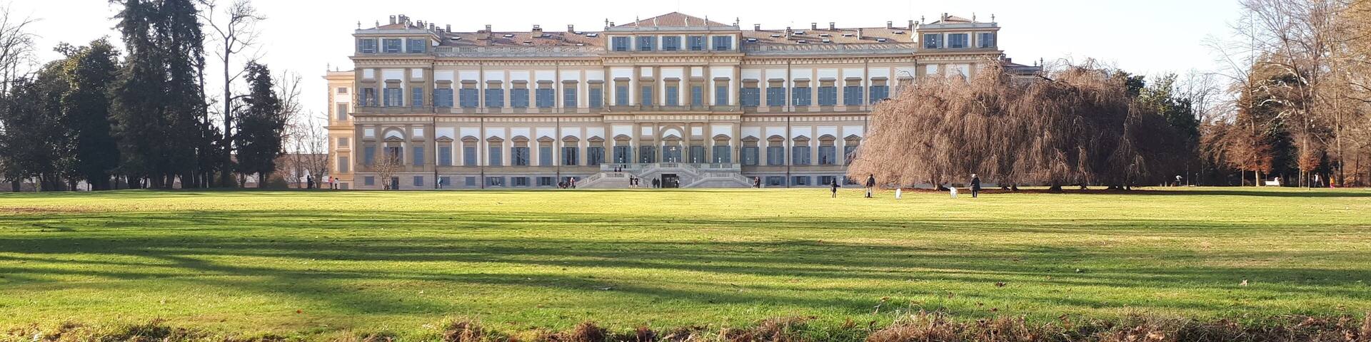 A large classical building with a symmetrical facade and a central staircase, surrounded by a well-maintained park with a pond in the foreground.