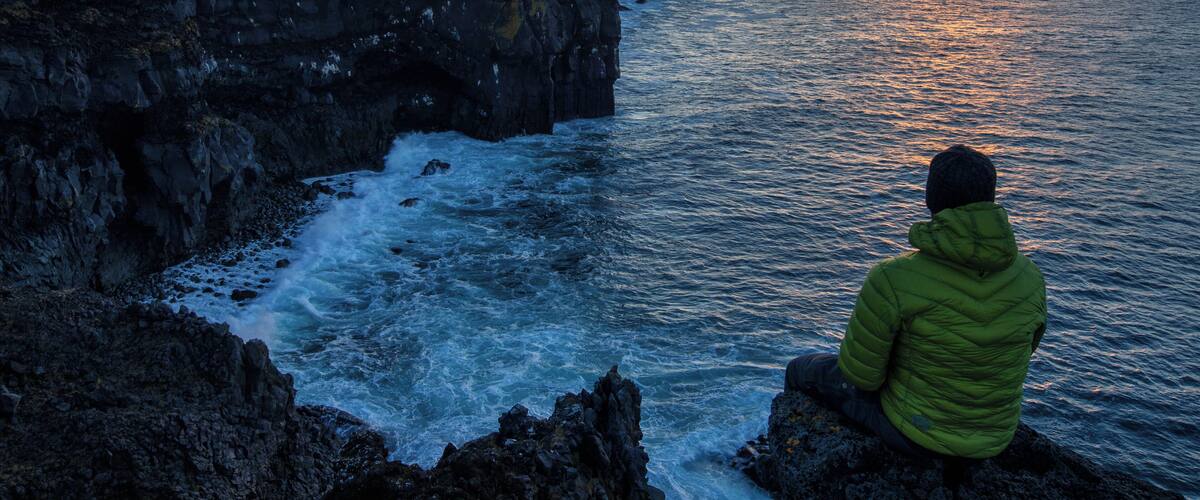 A person in a green jacket sitting on a rocky outcrop overlooking a coastal landscape during sunset.