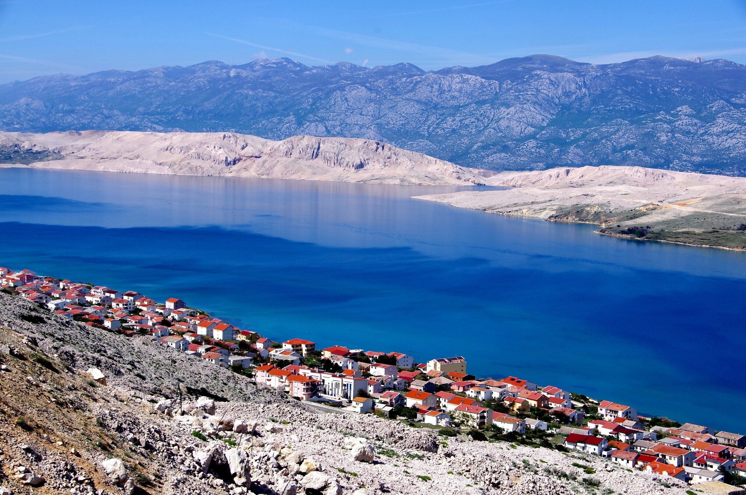 A coastal town with red-roofed buildings overlooking a large body of water and mountains in the background.