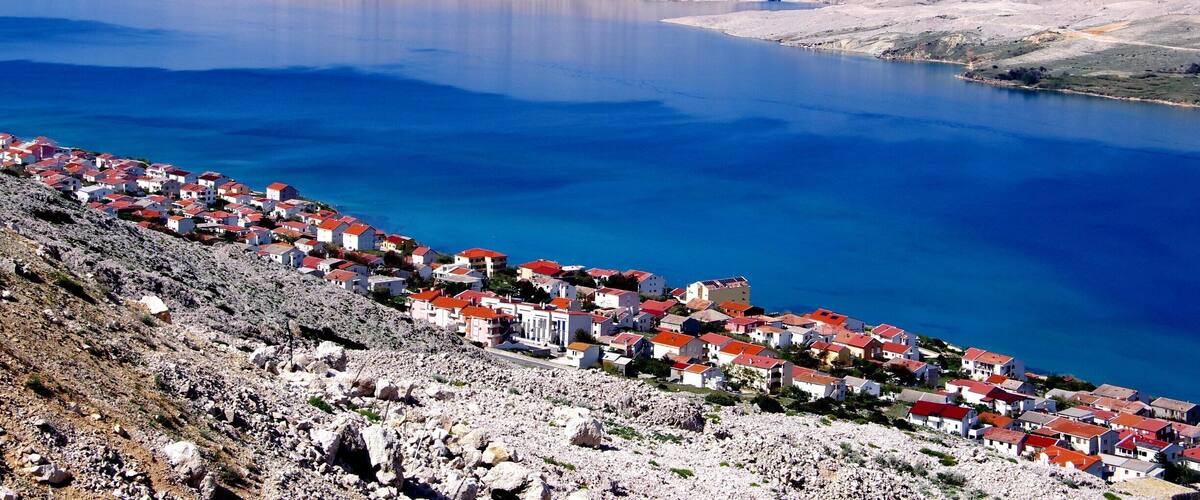 A coastal town with red-roofed buildings overlooking a large body of water and mountains in the background.