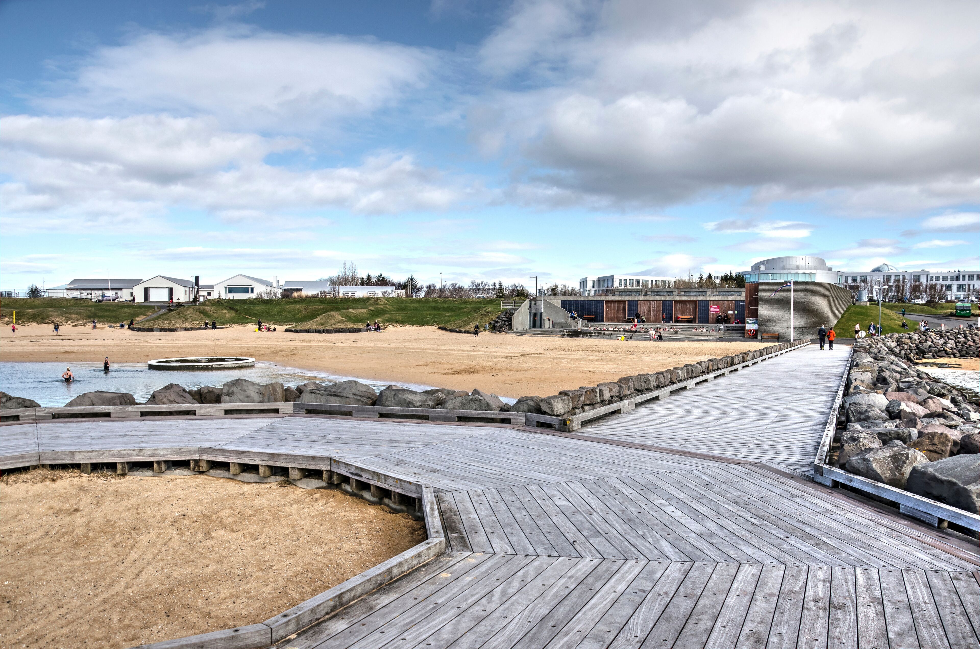 Un paseo marítimo de madera que lleva a una playa de arena con costa rocosa, un edificio moderno con cúpula, y un cielo despejado.