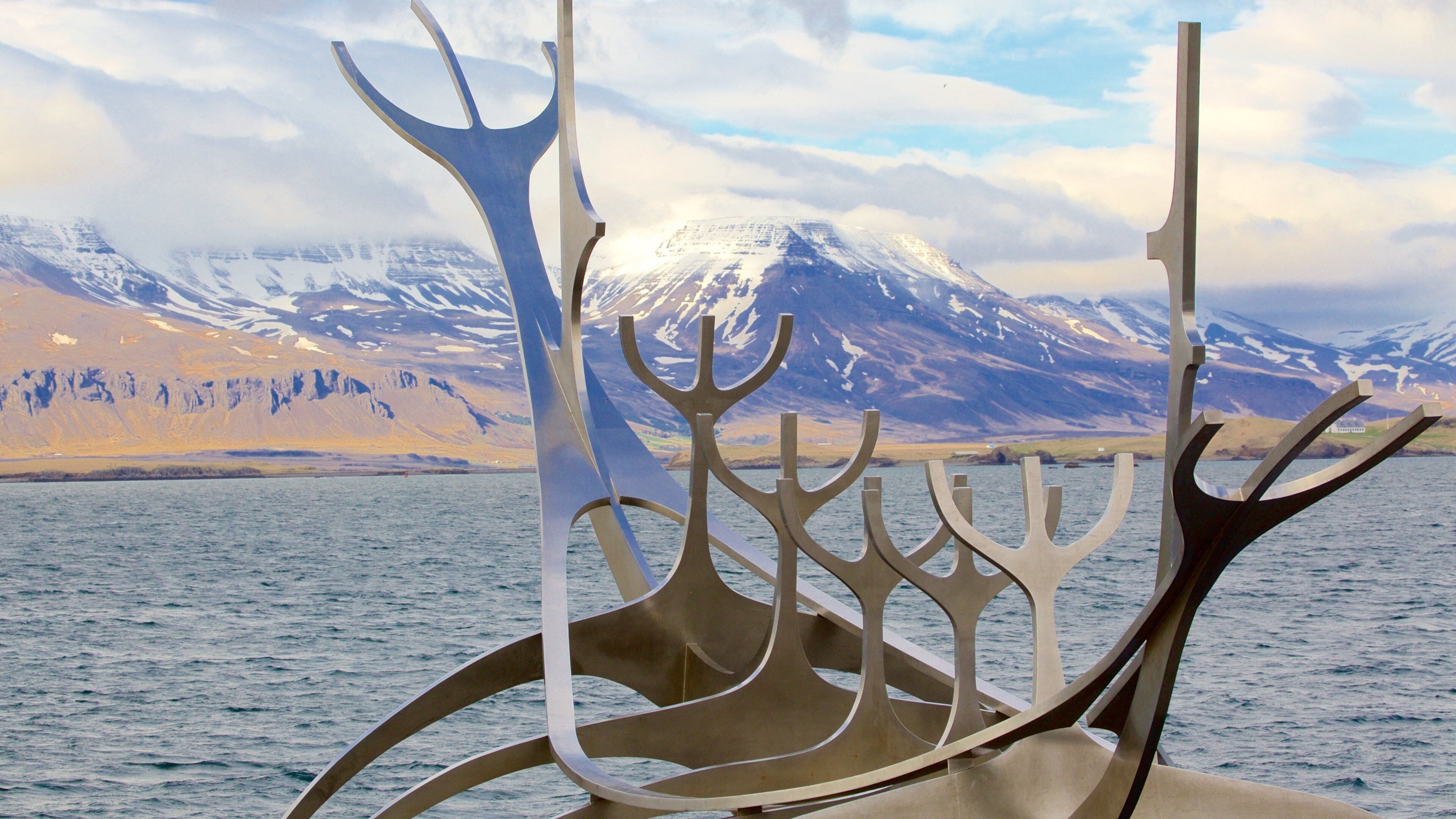 Une sculpture métallique se dressant devant un paysage montagneux, avec un plan d’eau au premier plan.