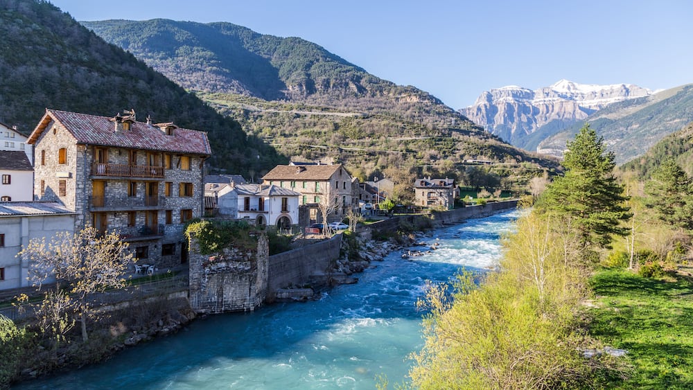 Une rivière traverse une ville où les bâtiments sont en pierre, avec des montagnes en arrière-plan.